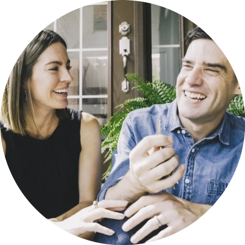 A laughing couple sits close together on the stoop of their new house. The person on the right holds the key to their house.